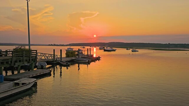 Peaceful marina at sunset with small boats, wooden docks, orange sky, reflective water, and distant shoreline creating a calm New England coastal travel scene.
