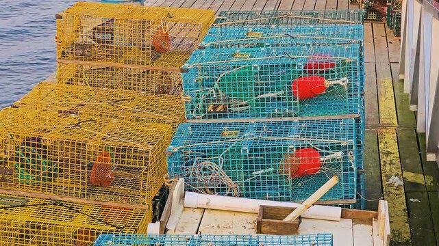 Large pile of lobster traps sits on a dock beside the harbor, with pink buoys, rope coils, weathered cages, and coastal homes across the water in a fishing town.