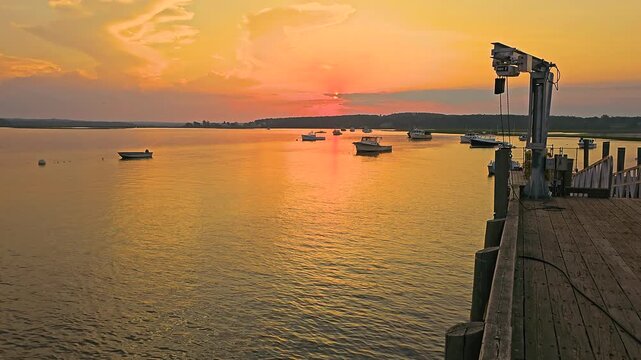 Beautiful waterfront sunset over a boat harbor, with warm orange light, still water, marina docks, moored vessels, and peaceful summer evening atmosphere by the coast.