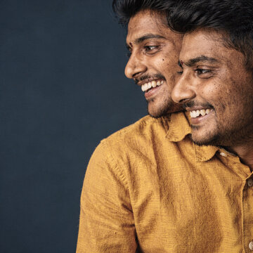 Two cheerful young Indian brothers smiling and looking away together against a dark blue background