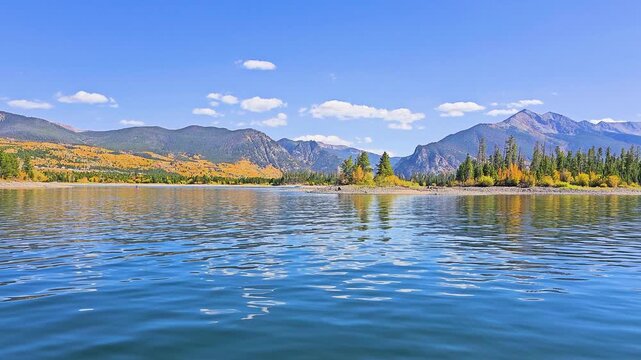 Boat level view of a mountain lake in fall, with gentle ripples, bright aspen reflections, evergreen shoreline, distant ridges, and crisp blue sky for scenic outdoor travel footage.