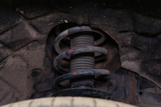 Dirty car suspension coil spring sits inside wheel well, with worn strut and dusty liner around it. Close detail reveals grime, rust specks