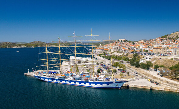 Aerial view of &Scaron;ibenik, Croatia, featuring a majestic tall ship moored in the harbor, with the historic coastal city, red rooftops, blue Adriatic Sea and Mediterranean landscape in the background.