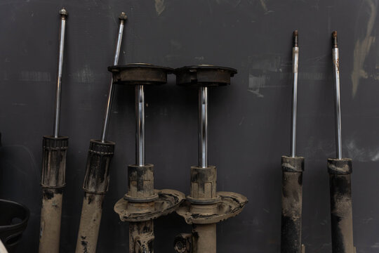 Used suspension struts and metal components rest in workshop storage against dark wall. Rust, grime, and scratched finishes suggest removal