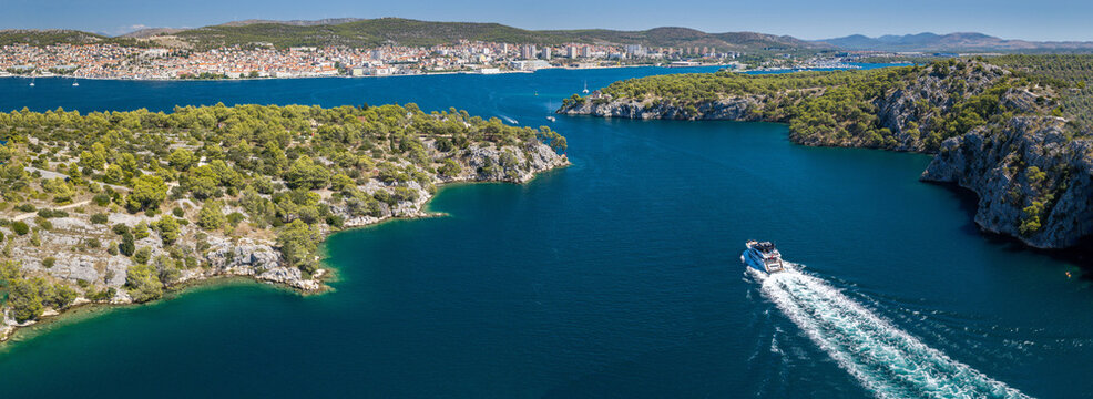 Aerial view of &Scaron;ibenik, Croatia, with a luxury yacht cruising through a scenic Adriatic channel, rugged pine-covered coastline, blue waters and the city skyline in the background.