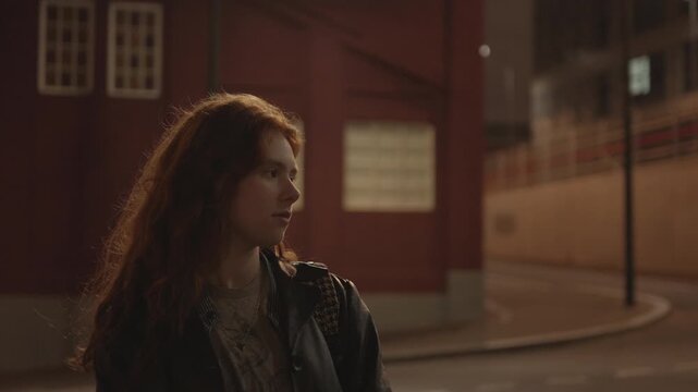 Cinematic night portrait of a young woman with long auburn hair wearing a leather jacket and backpack, standing on a quiet city street. Warm street lighting creates a moody, contemplative atmosphere 