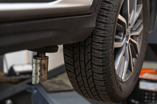 Automobile tire raised for service reveals tread, rim, and suspension area in garage. Metal lift and jack hold wheel steady, showing maintenance