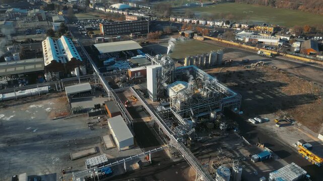 Industrial complex sprawls across vast, sunlit terrain. Smoke drifts from towers amid metal structures and pipes. Dark gritty chemical plant. Producing pollution and adding to global warming