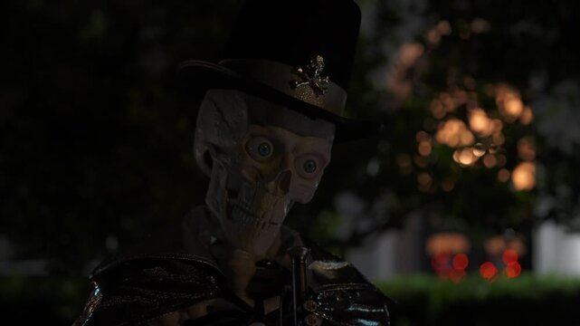 Close-up Halloween skeleton with bright glowing eyes, a top hat, and a dark cape. Bokeh lights and a suburban yard at night create a spooky festive scene.
