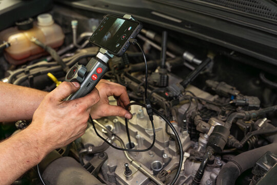 Mechanic work in engine bay with hands holding handheld diagnostic camera near wiring and hoses. Car repair task focuses on close access to hard to reach components and careful visual checking.
