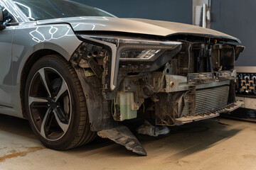 Car front corner rests in workshop with silver body panel, exposed headlamp housing, and missing bumper cover. Dusty parts and open structure reveal mechanical repair need during bodywork © Egor Lint