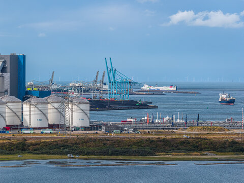 Antwerpen, Belgium - 16 August 2026: Aerial view of the Port of Antwerp-Bruges industrial area with storage tanks, shipping cranes, and cargo ships in the waterway under a blue sky.