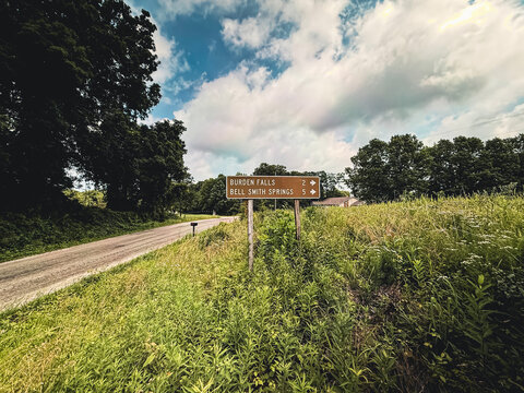 Wide perspective of a brown directional road sign to Burden Falls and Bell Smith Springs, set against grassy roadside foliage and blue sky in Shawnee National Forest, IL.