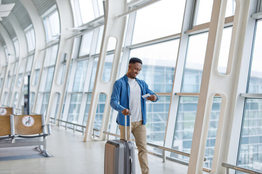 A smiling man in casual attire walks through a bright airport concourse with his luggage and travel documents in hand