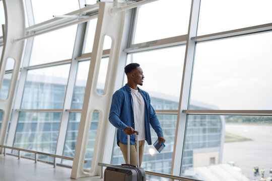 A young Black man stands by a large window in an airport terminal, holding his passport and ready for his flight