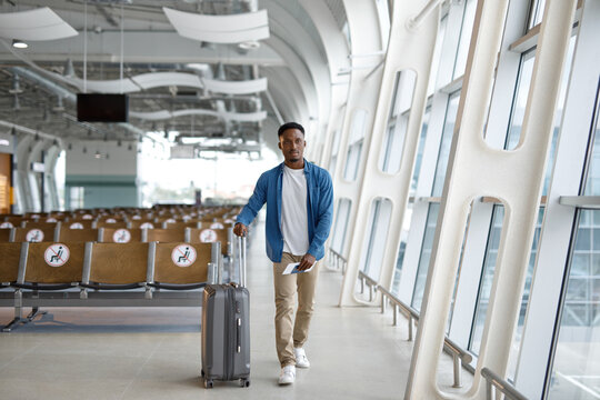 A young Black man pulls his luggage through a contemporary airport concourse, holding his travel documents, with empty seats marked for distancing