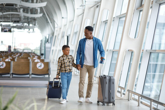 A father and his young son walk together through a modern airport terminal, pulling their suitcases and holding hands