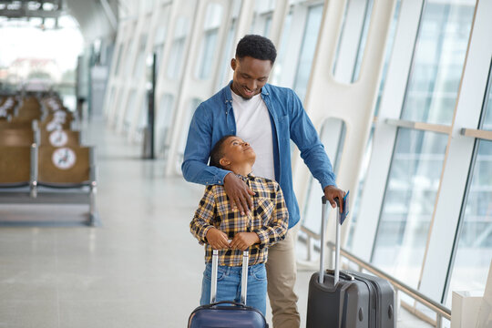 A father and his young son stand together in a modern airport terminal, looking at each other with smiles while holding their travel bags