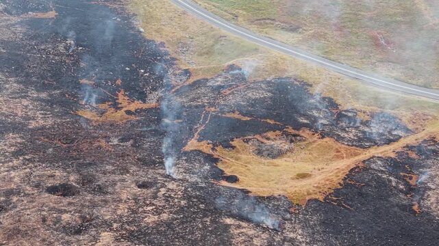 Wisps of smoke drifting from smouldering peatland post moorland fire, charred heather and blackened vegetation covering hill slope