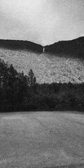 Fototapeta premium Distant Narrow Waterfall Between Forested Hills in Heavy Grain Black and White Landscape