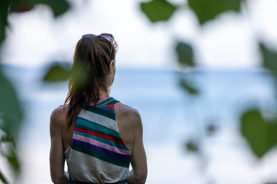 A brown-haired woman with her hair tied in a ponytail, surrounded by greenery, looking out over the sea.