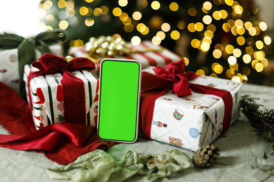 Smartphone mockup with green chroma key screen standing near wrapped Christmas gift boxes. Warm bokeh lights from Christmas tree. Concept of holiday e-commerce, mobile shopping and tech gifts.