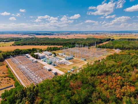 High angle shot of electrical power grid station near autumn woods. Industrial substation with metal pylons next to green and orange forest under sunny sky. Aerial perspective.