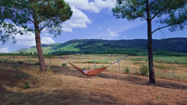 Young man relaxing in hammock with mountain view
Wide outdoor view of a young man gently swinging in a hammock while using his smartphone, surrounded by green mountains, trees and clouds.