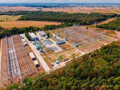 Top view of electrical distribution substation surrounded by fields. High voltage transformer station with steel pylons, control buildings and nearby woodland. Aerial shot.
