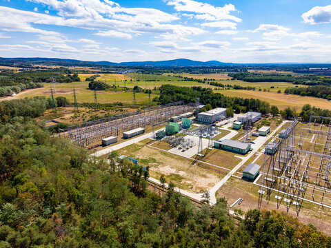 Aerial overview of an electrical substation and surrounding fields. Steel switchyard structures stand near green forest and agricultural land under a scattered cloud sky.