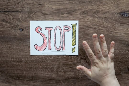 Birds eye flat lay photo of real human hand and Stop! handwritten on white sign.