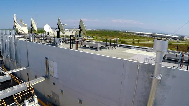 Aerial drone shot of industrial rooftop featuring multiple satellite dishes, communication equipment, metal structures and utility systems, with open green land and distant buildings in background