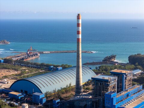 Aerial view of a power plant with a tall striped chimney, storage warehouse, and industrial facilities along the coastline with a cargo ship in the harbor in Pacitan, East Java, Indonesia.