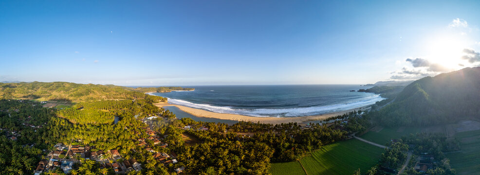 Aerial view of a coastline featuring a sandy beach, rolling waves, lush palm forests, and green fields under a clear blue sky in Pacitan, East Java, Indonesia.