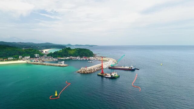 Aerial pullback from Jukdo Beach revealing fishing harbor breakwater construction with crane barge placing tetrapods beside turquoise coastline