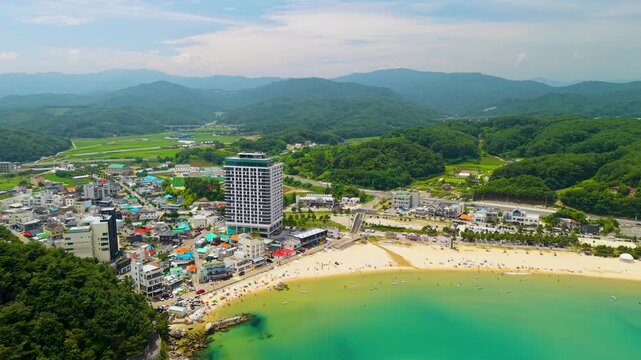 Aerial parallax over Jukdo Beach crowded with summer tourists in turquoise water beside coastal town of Hyeonnam-myeon with Gangwon-do mountains behind