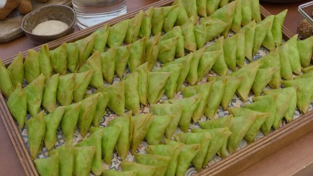 Triangular green pastries lined in rows on a tiled tray beside a dipping sauce bowl