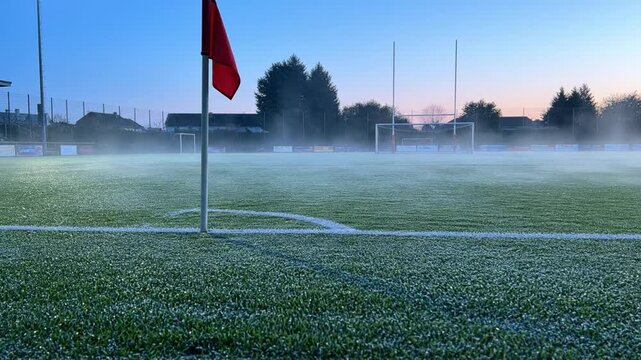 A red corner flag stands on a frozen soccer pitch at dawn with early morning mist blanketing the green grass in an open field.