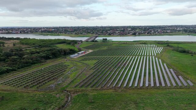 Solar panels form long rows beside Arroyo Martires, with Chacabuco Bridge, wooded banks and the Parana River visible beyond in Posadas, Misiones, Argentina, drone forward flyover.