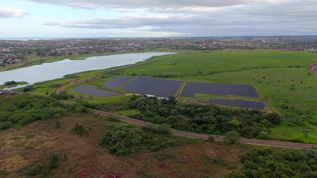 Photovoltaic panels extend beside Arroyo Martires in western Posadas, with green fields, wetlands and urban development beyond, drone forward flyover.