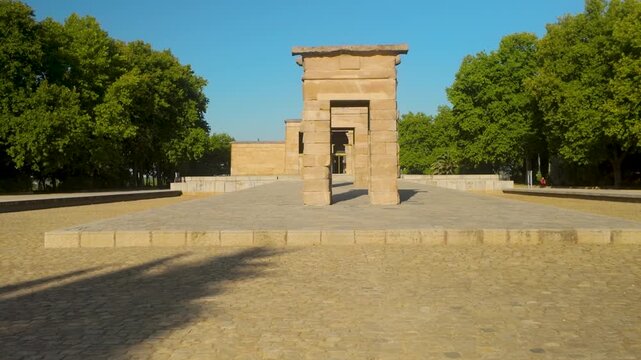 A wide shot of the Temple of Debod, an ancient Egyptian temple relocated to Madrid, surrounded by lush green trees under a clear blue sky.