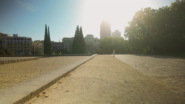 A wide angle view of a stone plaza in Madrid, Spain, featuring lush trees and city buildings under a bright setting sun. Captures urban wellness and authentic travel vibes.