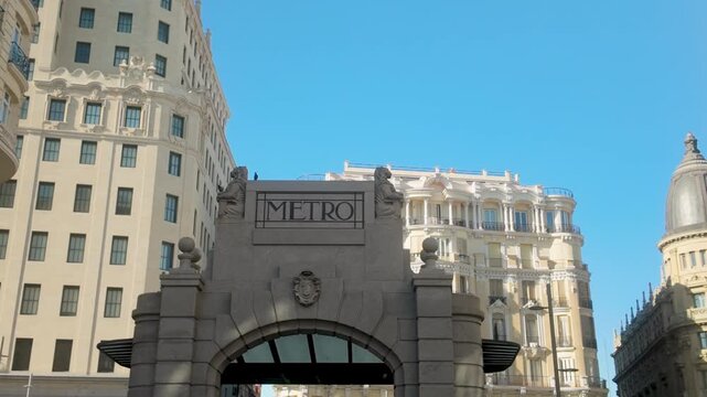 A low angle shot of the iconic stone Metro entrance in Madrid, Spain, surrounded by grand historic buildings under a clear blue sky.