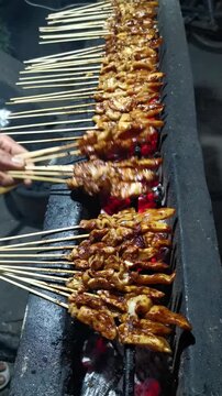Horizontal shot of a street vendor hand flipping chicken satay sate ayam skewers on a glowing charcoal grill at night