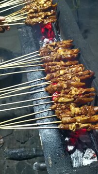 Vertical close up of grilling chicken satay sate ayam skewers over red hot charcoal embers at night