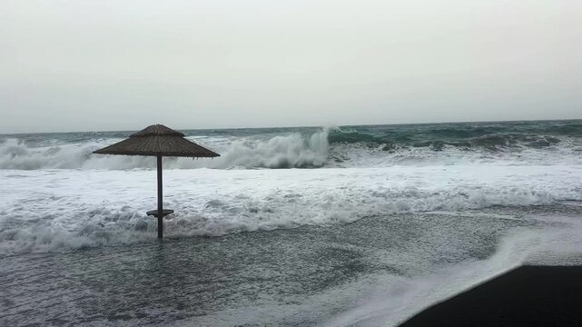 Rough waves crash across the dark volcanic shore at Vlychada Beach, Santorini, surrounding a lone straw umbrella with swirling foam and dramatic Aegean Sea surf.