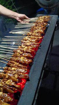 Vertical shot of street food vendor grilling rows of chicken satay sate ayam on hot charcoal embers at night