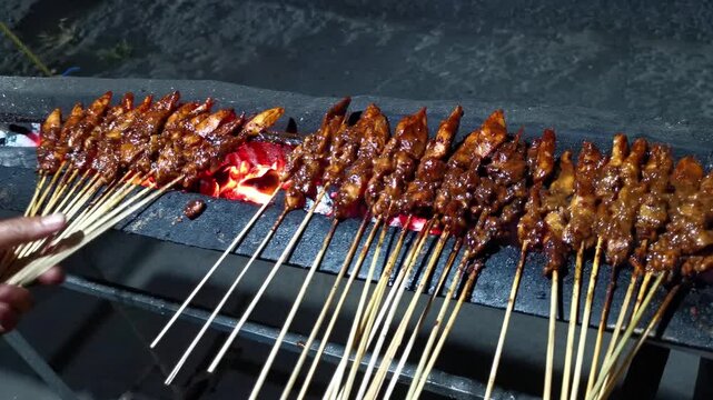Horizontal shot of a street vendor hand flipping chicken satay sate ayam skewers on a glowing charcoal grill at night