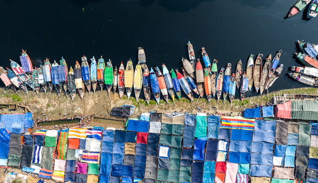Aerial view of colorful wooden boats docked along the riverbank next to a bustling market area with vibrant tarpaulin shelters in Tongi, Dhaka Division, Bangladesh.
