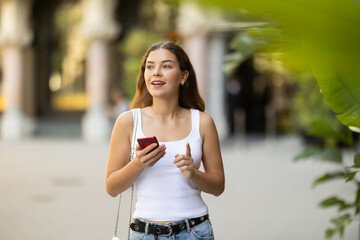 Young smiling girl with phone walks along city street, walks from work to home. Pedestrian in summer European capital, person in city, city bustle.